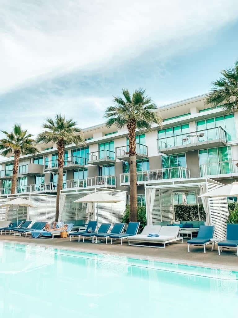 Luxury resort poolside with cabanas and loungers under tall palm trees, highlighting tranquil vacation vibes with a clear blue pool reflecting a bright, partly cloudy sky.