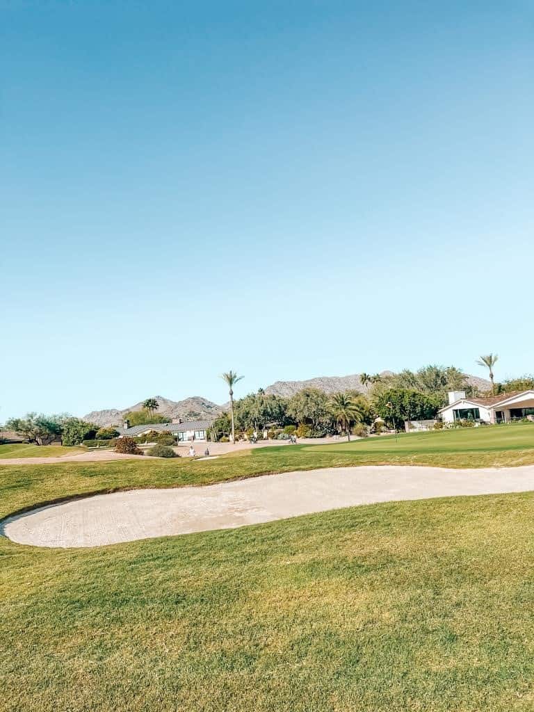Expansive view of the lush Mountain Shadows golf course with vibrant green fairways, a sand bunker in the foreground, and a backdrop of the picturesque Camelback Mountain under a clear azure sky.