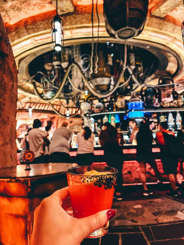 A hand holding a vibrant red cocktail with a rim of black salt, foregrounding the unique, dimly-lit interior of Oga's Cantina with patrons enjoying the immersive Star Wars-themed bar.