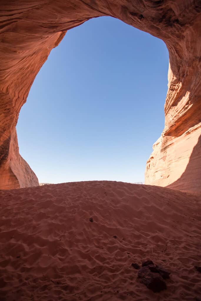 View from the base of a red sandstone arch in Page, Arizona, showcasing the smooth curves of the rock formation against a clear blue sky, with the sun casting shadows over the textured red sand.