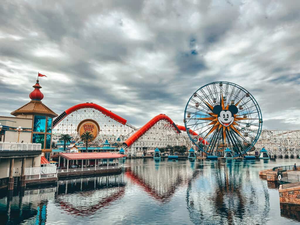 View of Pixar Pier at Disneyland Resort featuring the Incredicoaster and Pixar Pal-A-Round Ferris wheel, with Mickey Mouse's face, reflected in the calm waters below on an overcast day
