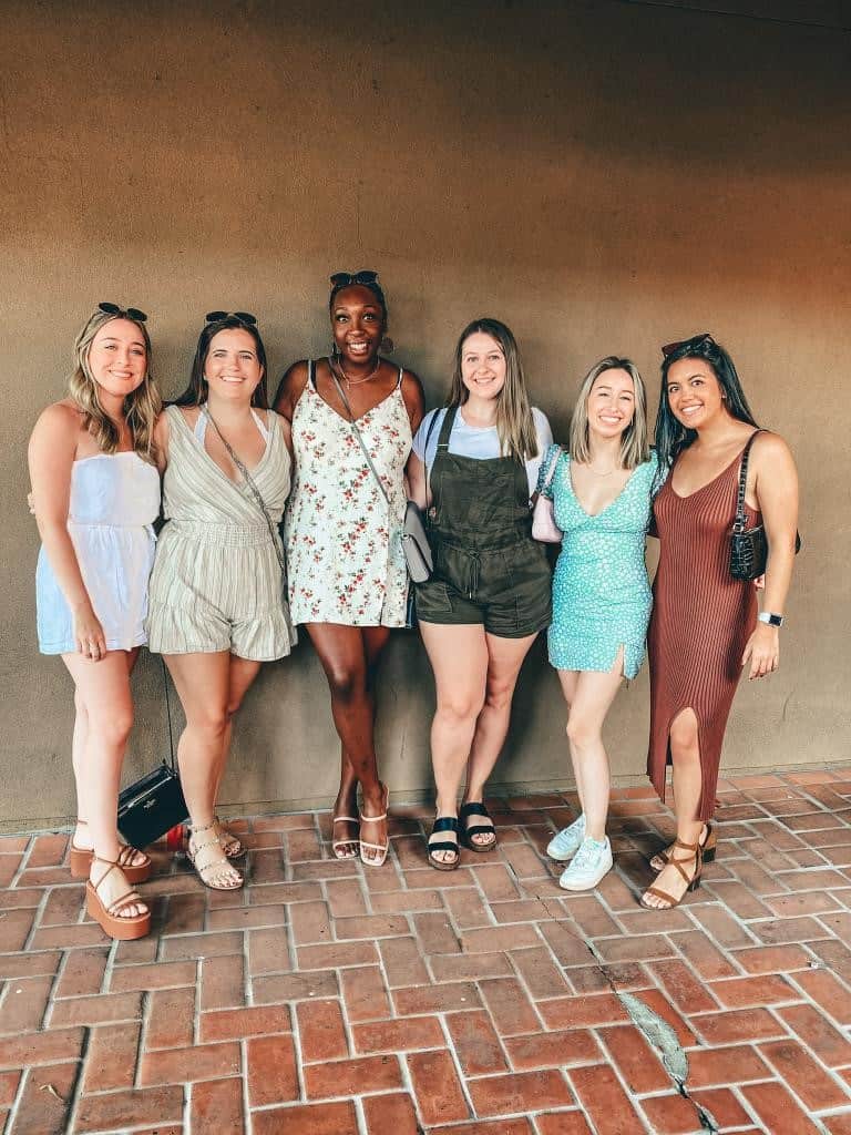 A group of six smiling women, friends enjoying a girls' trip in Scottsdale, pose for a photo against a textured adobe wall. They are stylishly dressed in summer outfits, ready to explore the city's charm.