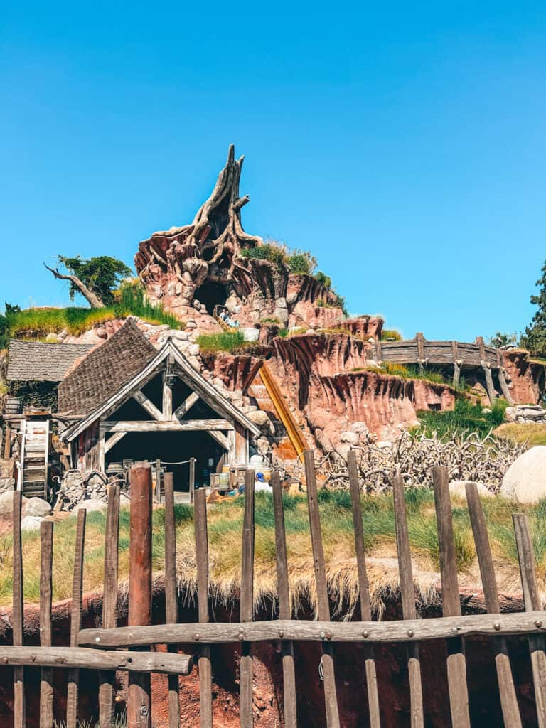 The iconic Splash Mountain, with its distinctive rocky and tree-lined peak, under a bright blue sky. A rustic wooden fence and cabin foreground add to the classic log flume ride's charming, wilderness-themed setting.