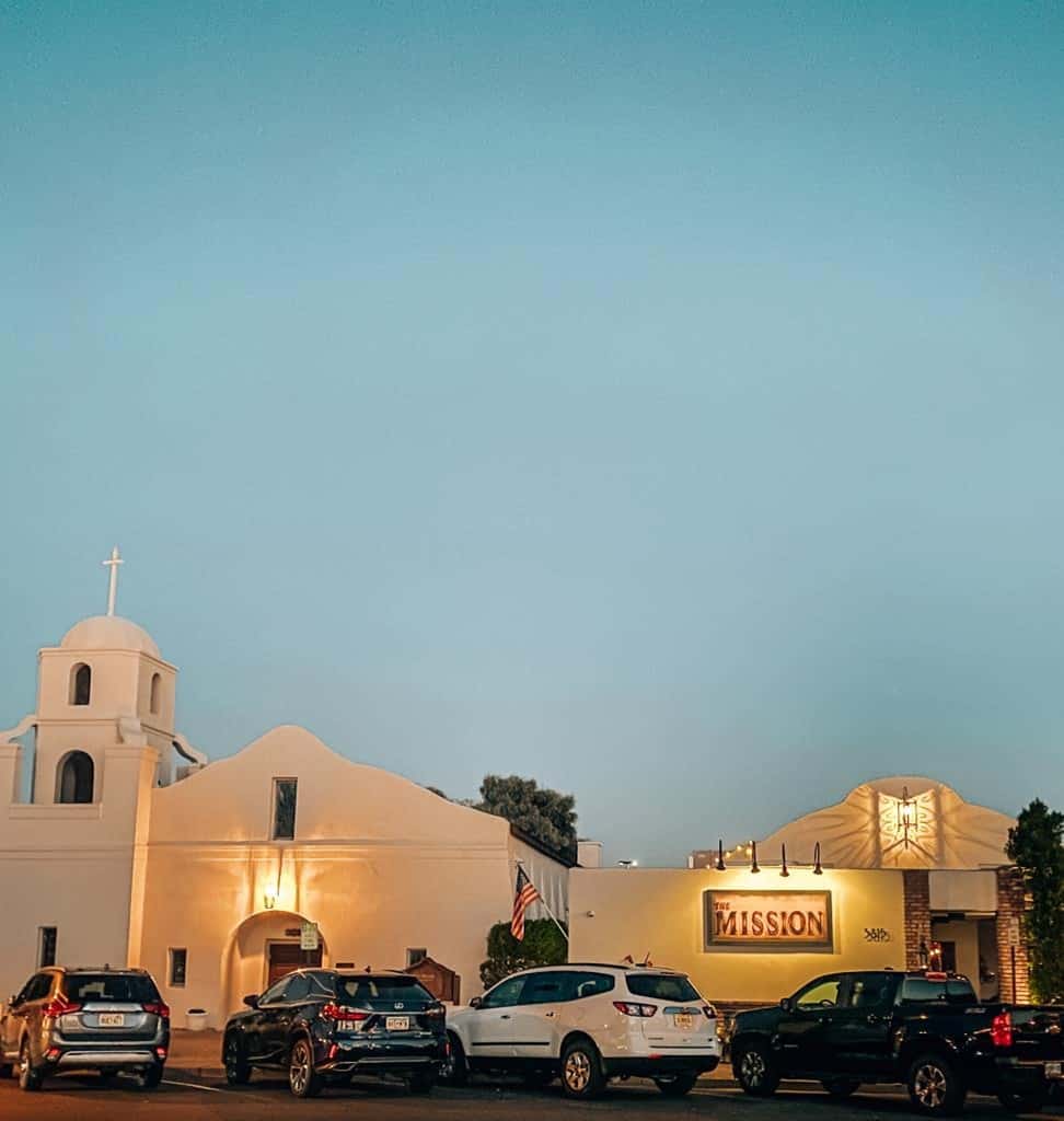 Twilight view of The Mission restaurant in Scottsdale, showcasing its iconic white mission-style architecture with a glowing sign, under a deep blue sky, embodying a blend of historic charm and modern dining ambiance.