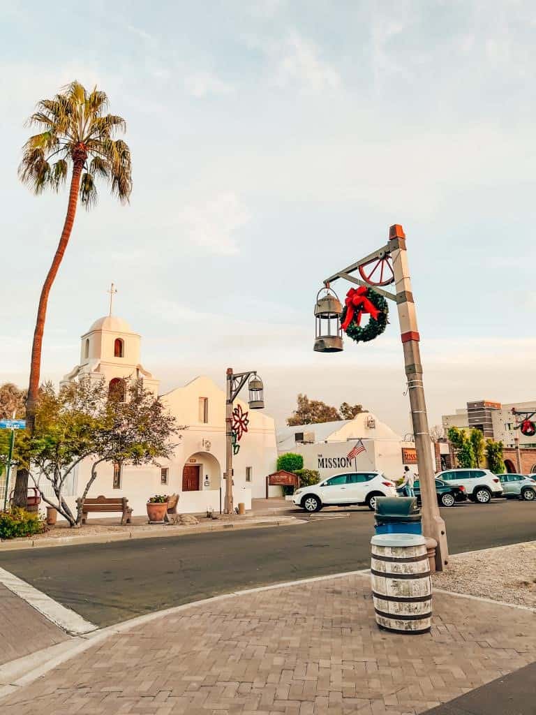 Charming street view at sunset with a historic white mission-style church, adorned with a Christmas wreath and festive decorations, next to a tall palm tree and cobblestone streets, evoking a warm holiday season in Scottsdale