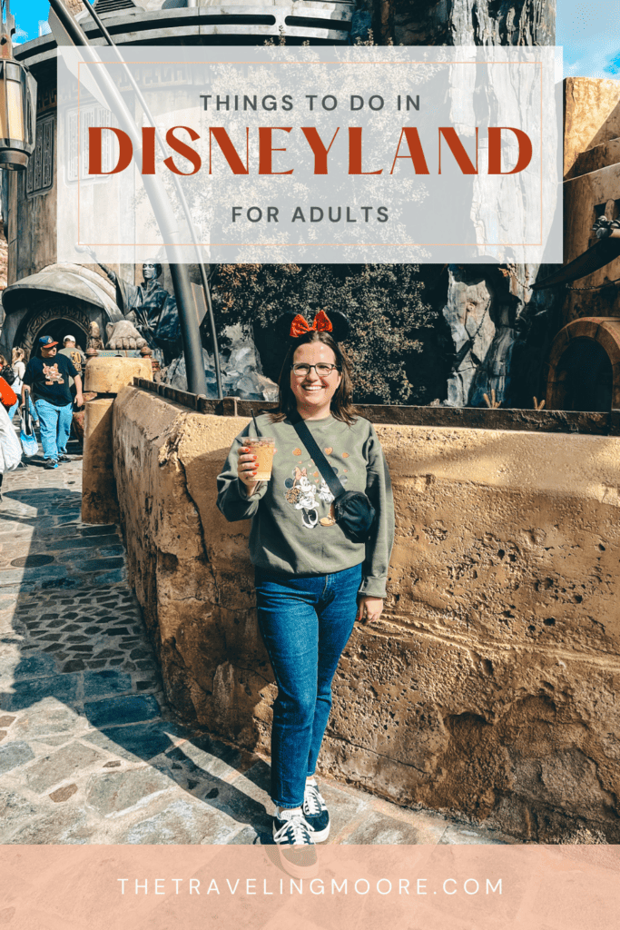 A cheerful adult woman holding a drink and wearing Mickey Mouse ears at Disneyland, with the text overlay 'THINGS TO DO IN DISNEYLAND FOR ADULTS' and the website 'THETRAVELINGMOORE.COM'. The backdrop features the immersive, rocky architecture of the park.