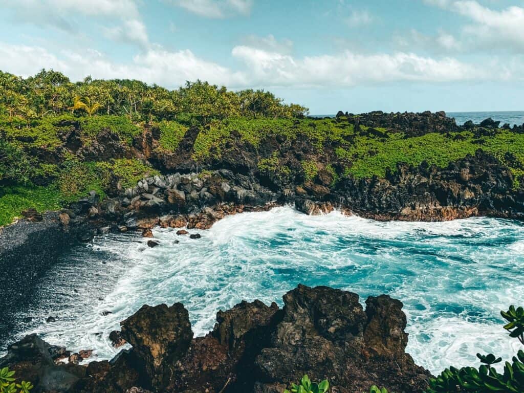 Vibrant tropical greenery fringes the rocky cliffs leading down to a secluded black sand beach at Waiʻanapanapa State Park on the Road to Hana, Maui, as frothy waves crash against the shore under a blue sky with scattered clouds.