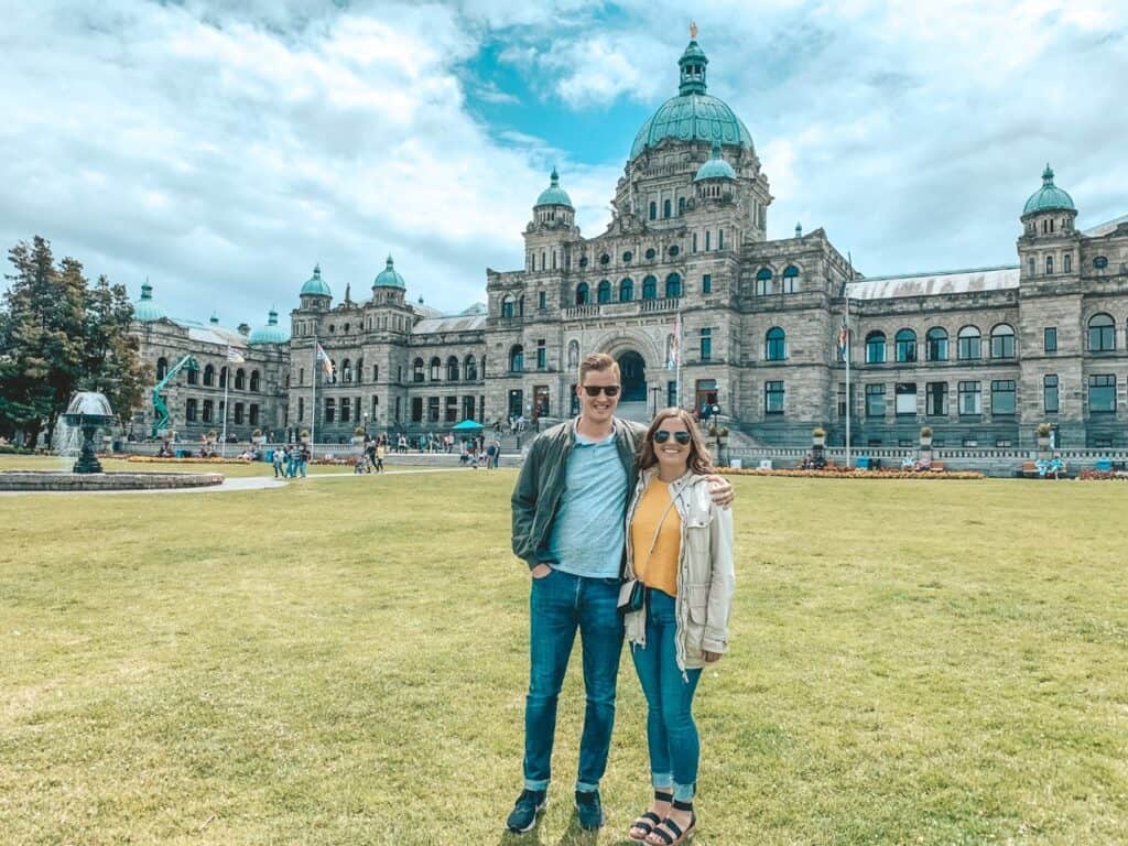 A smiling couple posing in front of the British Columbia Parliament Buildings in Victoria, capturing their travel memories against the grandiose and historic architecture