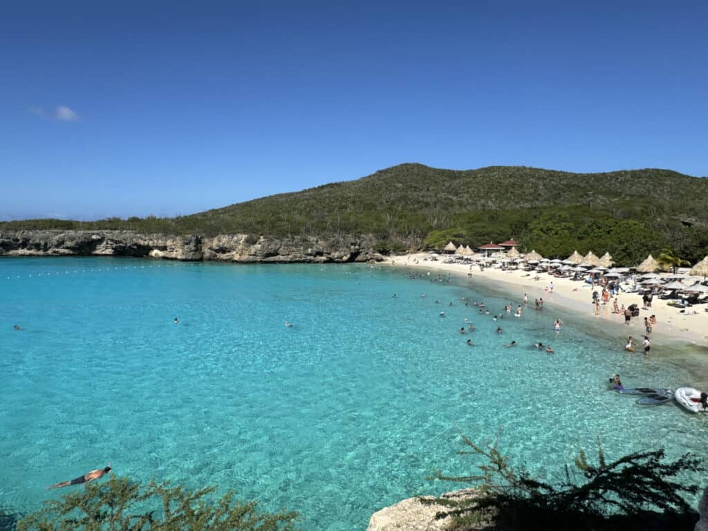 Grotto Bay Beach in Curacao, bustling with beachgoers enjoying the sun on the white sand and swimming in the crystal clear turquoise waters, with rugged cliffs and lush hills surrounding the bay under a brilliant blue sky.