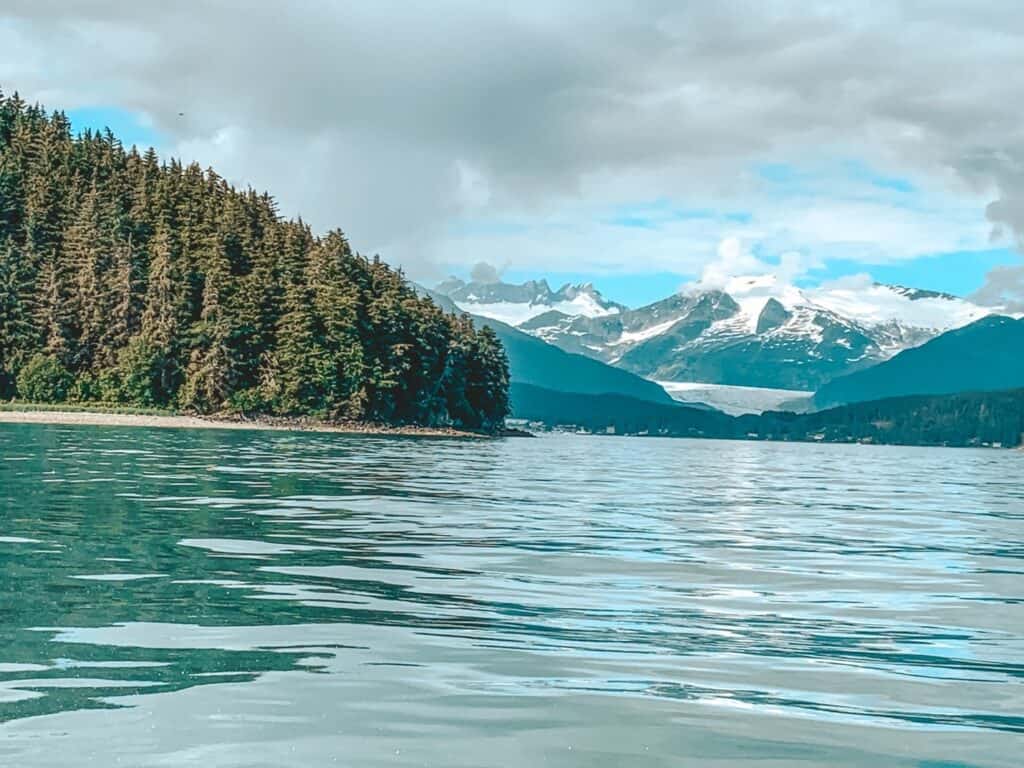 Scenic Alaskan landscape with lush pine forest on a coastline leading to snow-capped mountains and the Dawes Glacier in the distance, reflecting serenity and the wild beauty of Alaska – perfect for Instagram captions celebrating nature's majesty