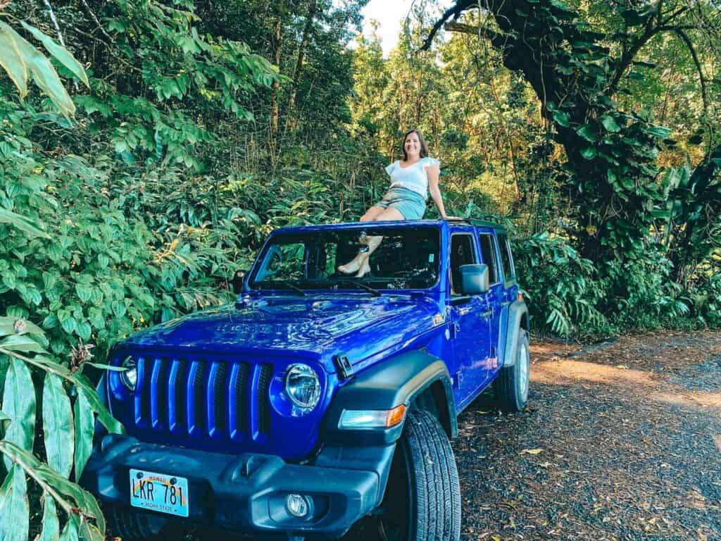 A woman sits atop a blue Jeep Wrangler parked on a dirt path surrounded by lush Hawaiian jungle foliage, embodying a sense of adventure on the Road to Hana