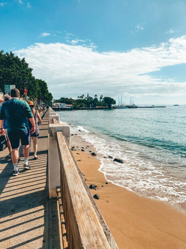 People strolling along the Lahaina boardwalk in Maui, with its rustic wooden railings, beside a calm sandy beach and clear waters, set against a backdrop of sailboats and tropical trees under a bright blue sky.