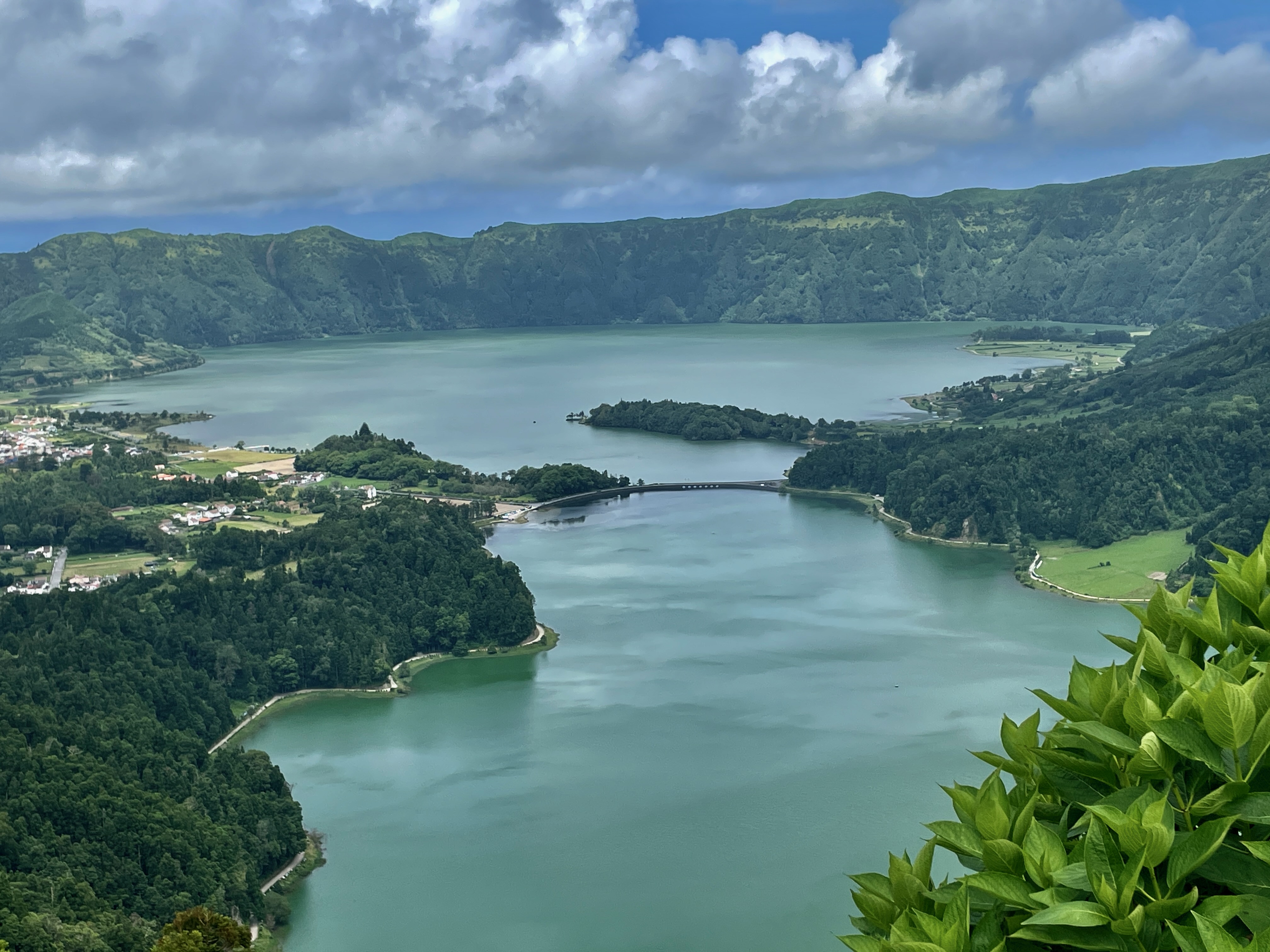 Aerial view of the Lagoa das Sete Cidades in São Miguel, Azores, showcasing the vibrant dual lakes separated by a bridge, surrounded by lush greenery and a quaint town under a cloudy sky.