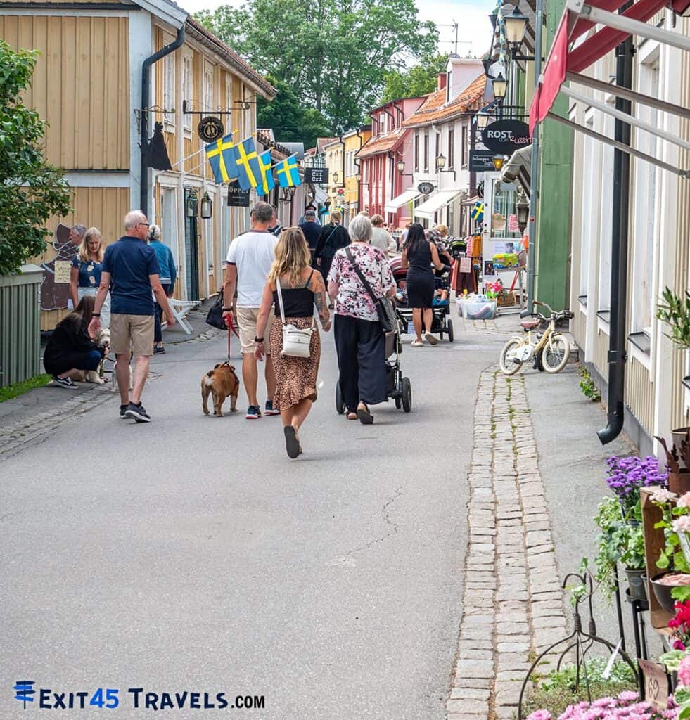 Busy street scene in an old town in Sweden, with people walking dogs and pushing strollers among colorful historic buildings, and a prominent Swedish flag fluttering in the breeze