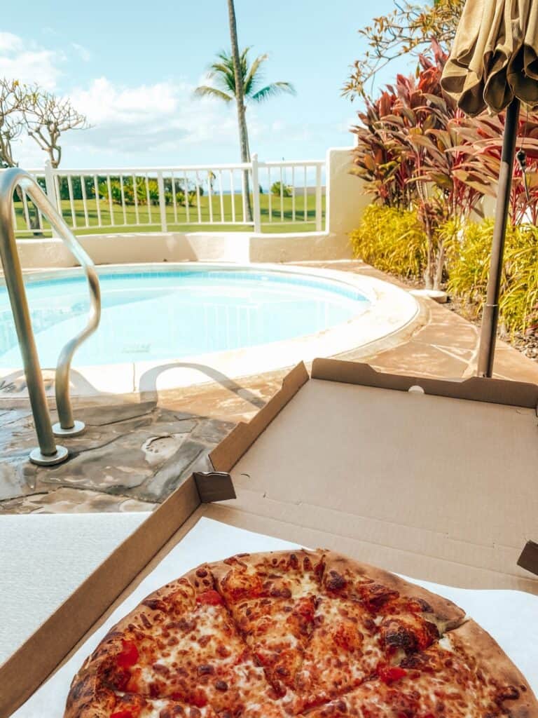 A fresh cheese pizza in an open cardboard box on a patio table beside a private plunge pool, with tropical foliage and a palm tree under a sunny blue sky, encapsulating a leisurely holiday vibe.