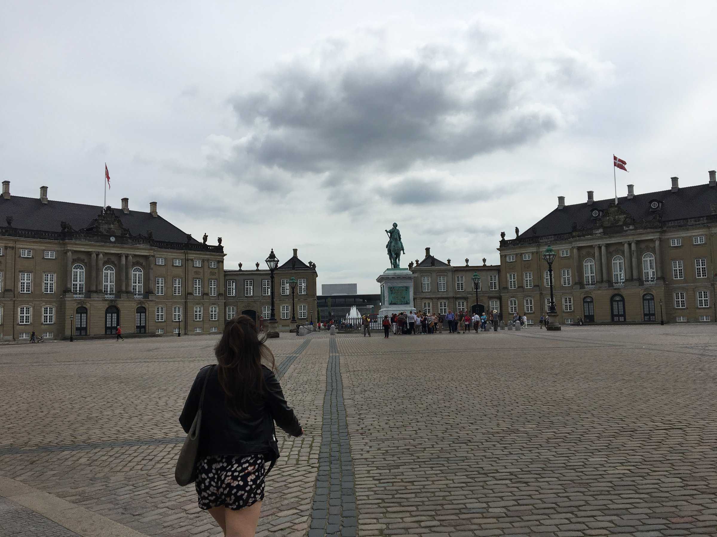 A woman walks towards the historic Amalienborg Palace in Copenhagen, Denmark, with its classical architecture and statue of King Frederik V on horseback in the center of the cobblestone courtyard, under a cloudy sky