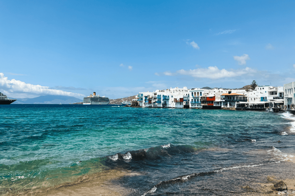 Vibrant Mykonos waterfront with a line of quaint, colorful buildings by the azure Aegean Sea, as cruise ships anchor in the distance, capturing the essence of Greek island life under a clear blue sky.