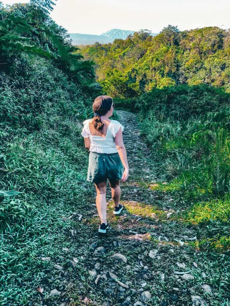 A woman hiking a rugged trail on the Road to Hana, surrounded by lush Hawaiian vegetation with a view of the distant mountains, capturing the essence of an adventurous trek through Maui's natural landscapes.