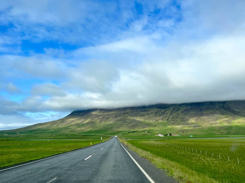 An open road in the Icelandic countryside during summer, leading towards a dramatic mountain partially covered with low clouds, with vibrant green fields on either side under a bright blue sky with scattered clouds