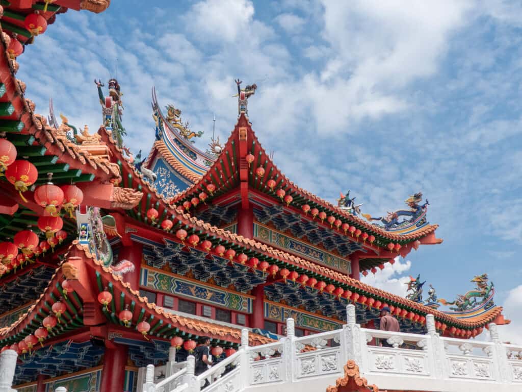 Ornate rooftop of a traditional Malaysian temple adorned with intricate designs, featuring vibrant red lanterns, detailed carvings, and mythological figures against a backdrop of a blue sky with wispy clouds