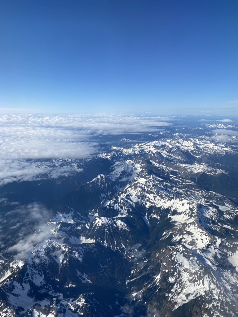 Aerial view of rugged mountain peaks with patches of snow under a clear blue sky, clouds carpeting the valleys between the ranges