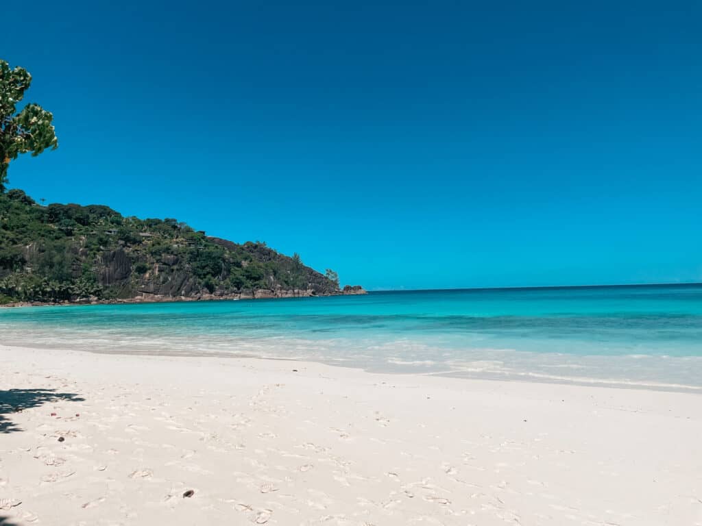 Idyllic view of a Seychelles beach with powdery white sand leading into crystal-clear turquoise waters, a lush green hillside in the background, under a vast, cloudless cerulean sky.