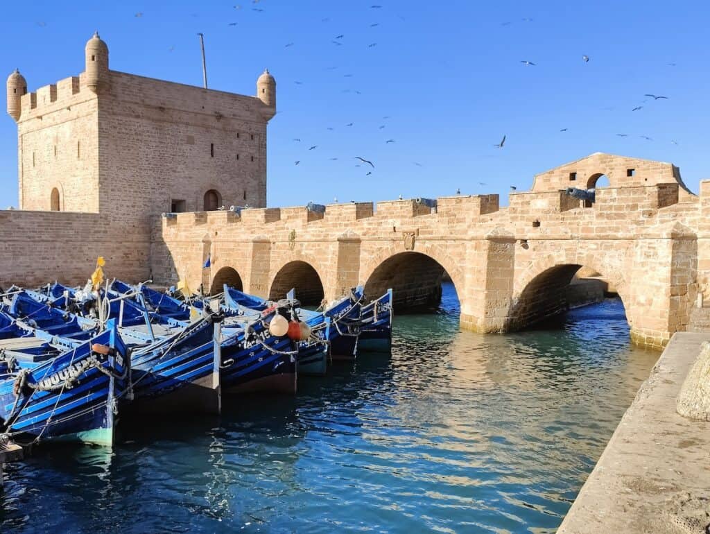 A picturesque view of the historic port in Essaouira, Morocco, featuring a stone fortress and arched bridge. Blue fishing boats are moored in the calm water, while seagulls soar in the clear blue sky above. The scene captures the charm and maritime heritage of this coastal town.