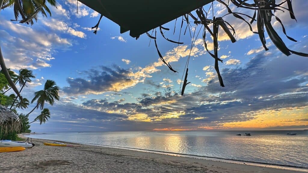 A tranquil beach scene in Fiji at sunset, featuring a sandy shoreline with a few kayaks and boats on the calm water. Palm trees frame the view, and the sky is filled with dramatic clouds lit by the setting sun, reflecting on the ocean's surface. The scene captures the serene beauty and tropical ambiance of this island paradise.