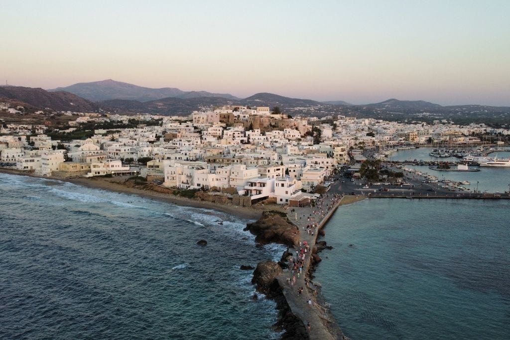 Aerial view of Naxos, Greece, at sunset, showcasing its whitewashed buildings clustered along the coastline and extending towards the hills. The calm waters of the Aegean Sea lap against the shore, and a long promenade leads out into the sea, bustling with people. The surrounding mountains create a picturesque backdrop for this charming island town.