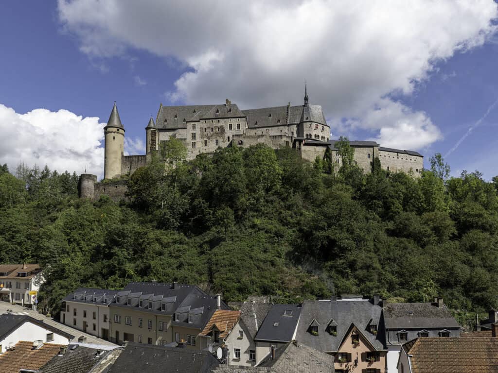 A majestic medieval castle sits atop a lush, green hill overlooking a quaint village in Luxembourg. The castle's towers and stone walls are prominent against a backdrop of a partly cloudy sky, while charming houses with dark slate roofs cluster at the base of the hill. The scene exudes a blend of historical grandeur and peaceful village life.