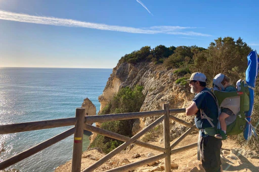 A man wearing a cap and sunglasses stands on a cliffside trail overlooking the ocean in Algarve, Portugal, carrying a child in a hiking backpack. The clear blue sky and calm sea create a serene backdrop, with rocky cliffs and greenery enhancing the scenic view.