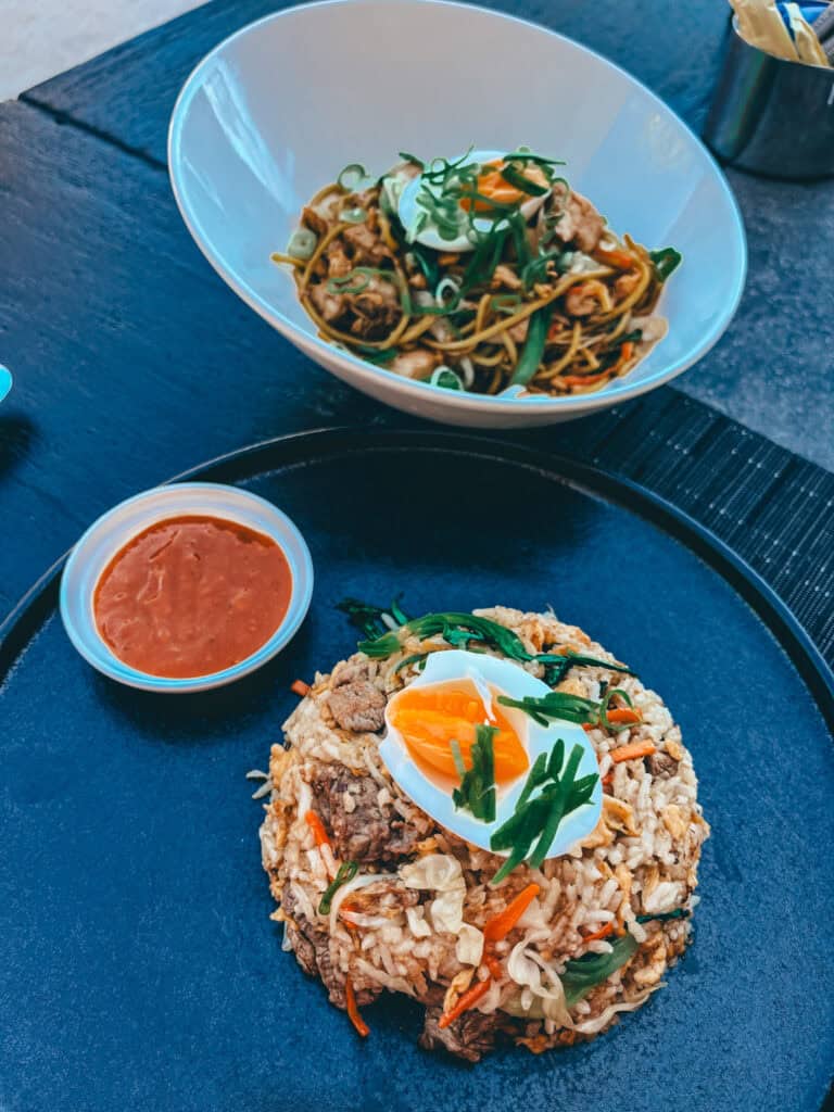 A plate of fried rice topped with a halved boiled egg and garnished with green onions, accompanied by a bowl of noodles with vegetables and meat, and a side of red sauce.