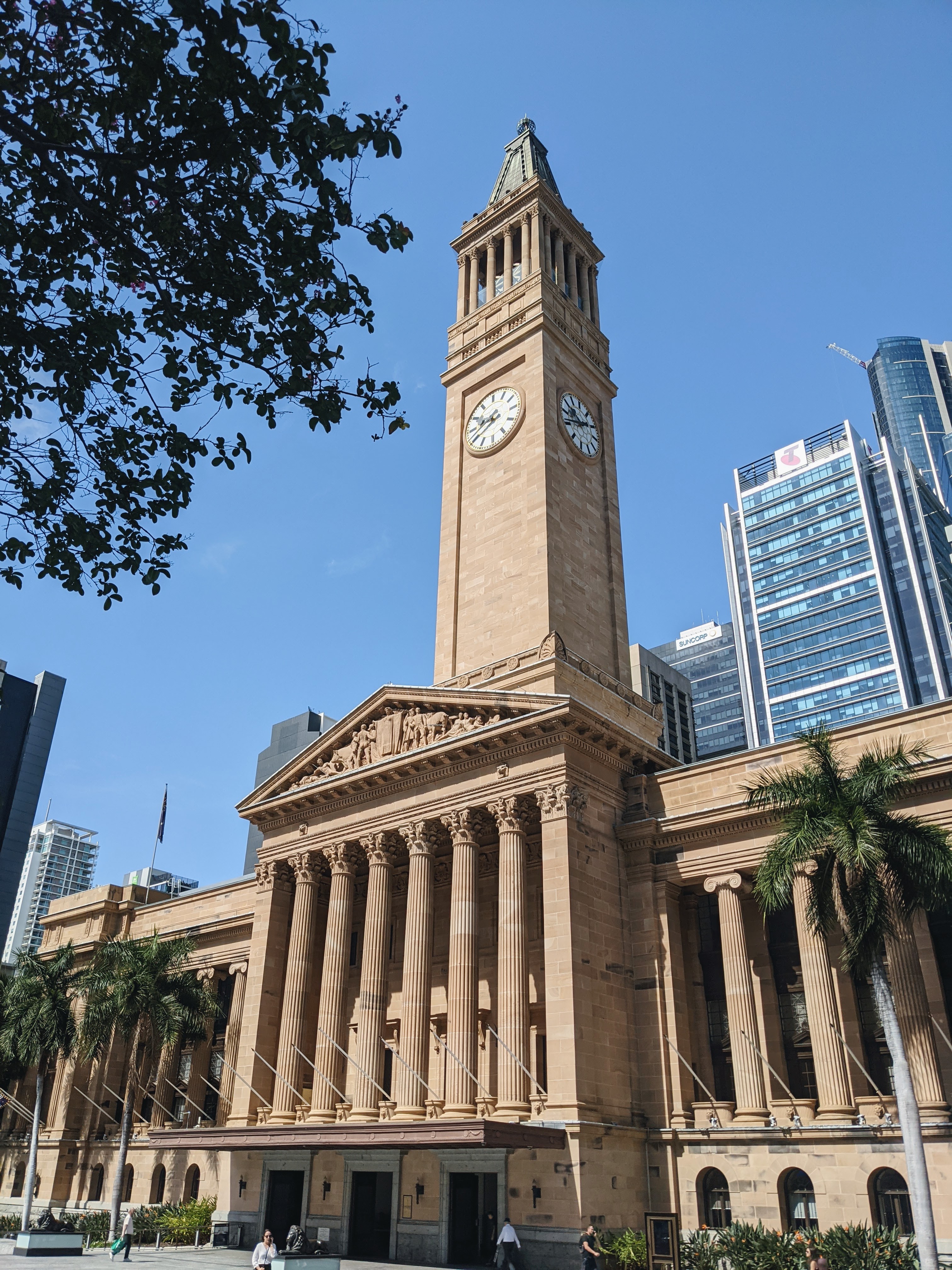 A view of Brisbane City Hall in Australia, featuring its prominent clock tower under a clear blue sky. The building showcases classical architecture with tall columns and detailed stonework, set against a backdrop of modern skyscrapers. Palm trees and a few people walking add to the vibrant urban atmosphere.