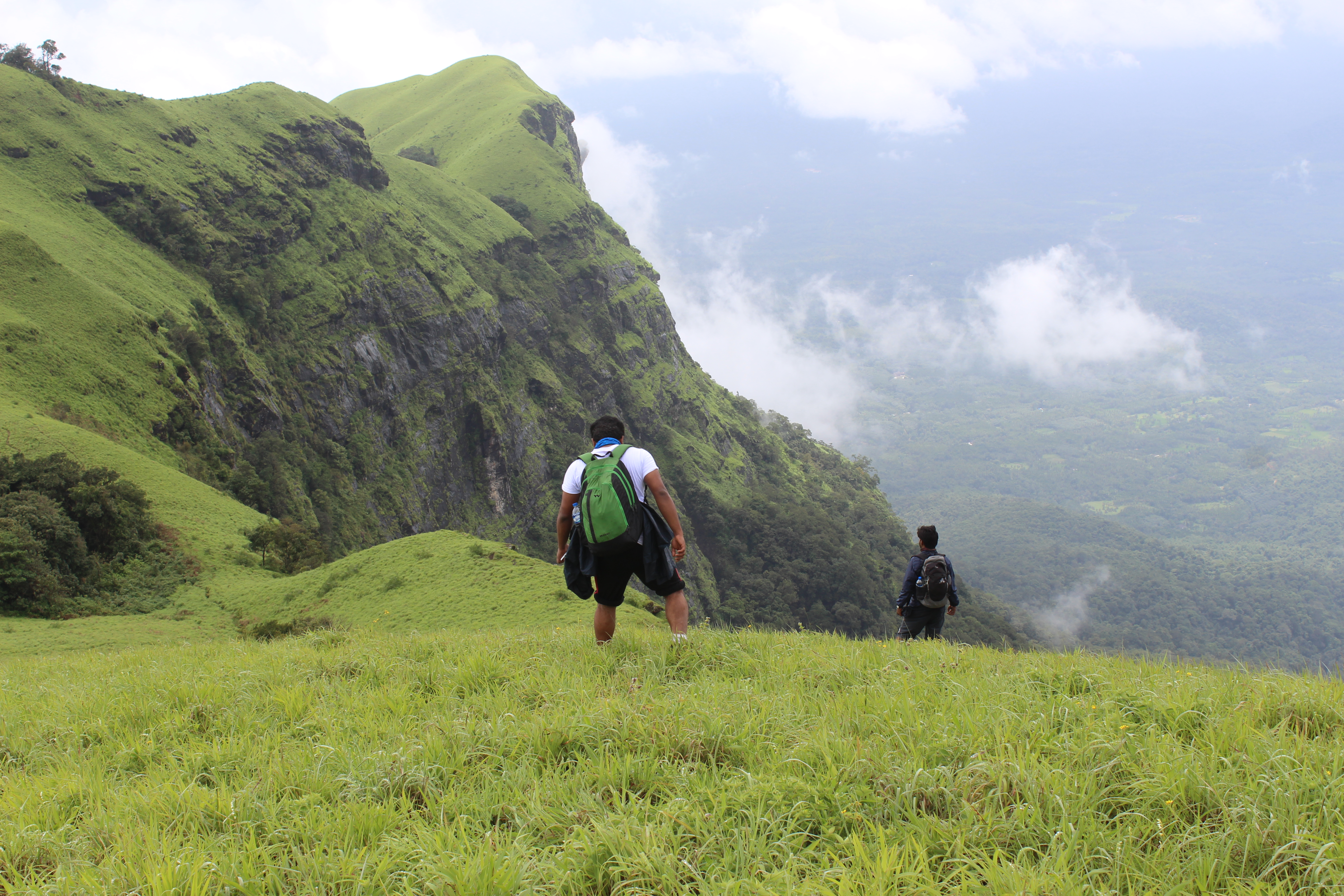 Two hikers traverse the lush, green hills of Ballarayana Durga in India, with a steep, rocky cliff in the background. The expansive view includes rolling hills and a misty valley below, creating a sense of adventure and natural beauty. The hikers' backpacks and gear add to the atmosphere of exploration in this scenic landscape.