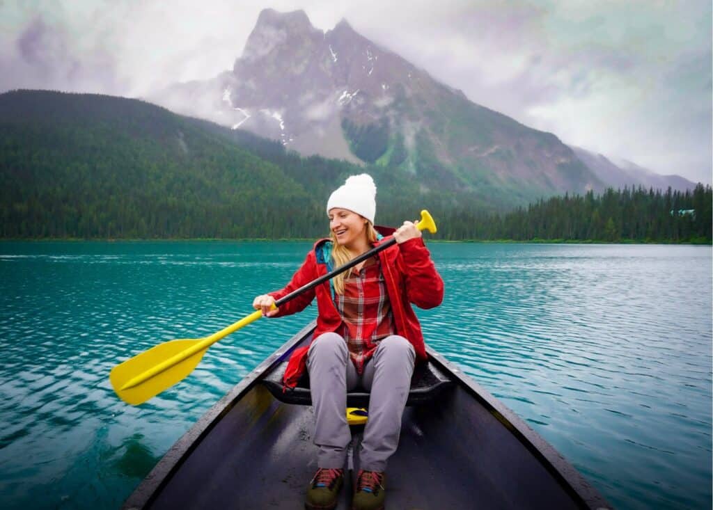 A cheerful woman paddles a canoe on a turquoise lake in Banff, Canada, surrounded by forested mountains and misty peaks. She wears a red jacket and a white beanie, enjoying the serene and picturesque natural setting. The vibrant colors and tranquil water highlight the beauty of this outdoor adventure.