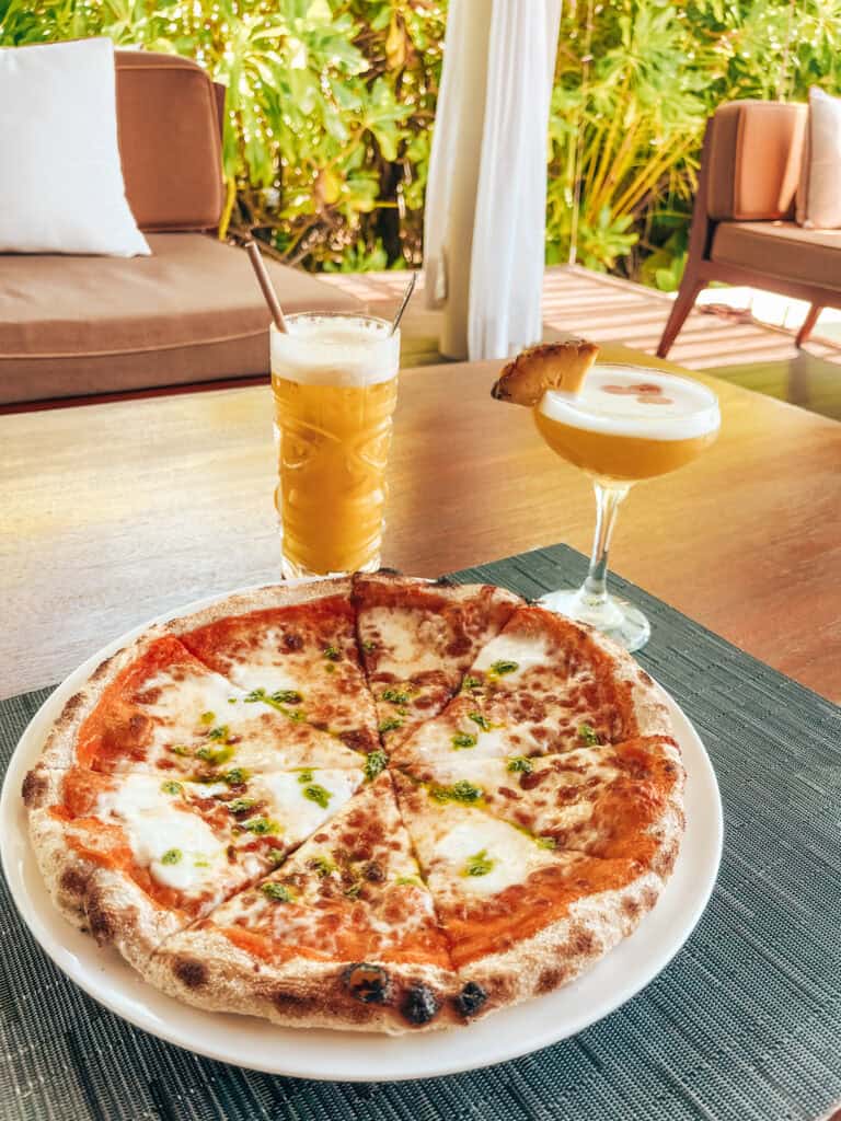 A wood-fired pizza topped with melted cheese and herbs, served with two tropical drinks: one garnished with a pineapple slice and the other a frothy cocktail, set on an outdoor table with lush greenery in the background.