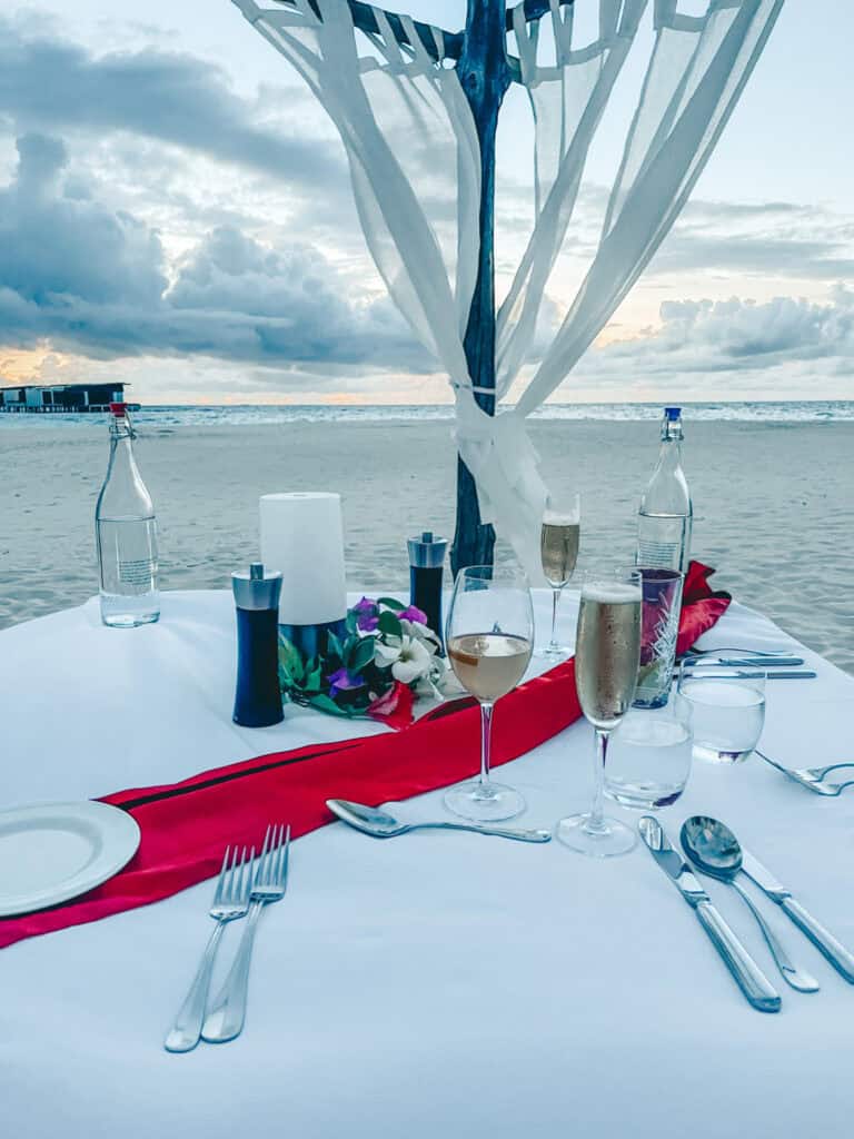 A romantic beach dinner setup featuring a table with a white tablecloth, red runner, and elegant place settings, surrounded by sheer white drapes and overlooking the ocean at sunset.