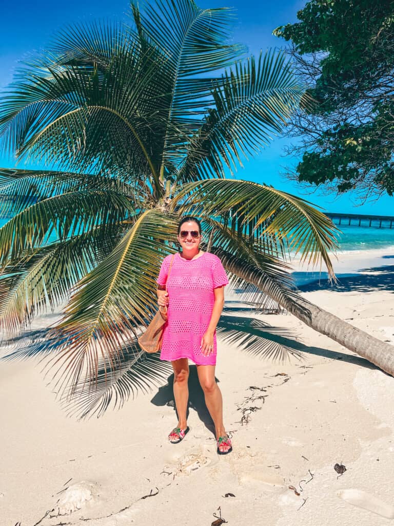 Woman in a bright pink crochet cover-up dress, floral sandals, and sunglasses, standing on a sandy beach with a palm tree and turquoise water in the background.