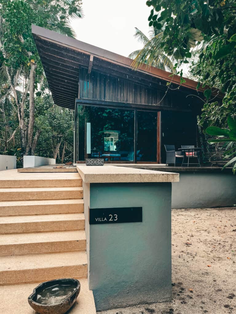 Exterior view of Villa 23 at the Park Hyatt Maldives, featuring a modern design with a slanted dark wooden roof and full-length glass windows. The villa is nestled among lush tropical greenery, with sandy ground leading to its entrance marked by sand-colored steps and a minimalistic sign.