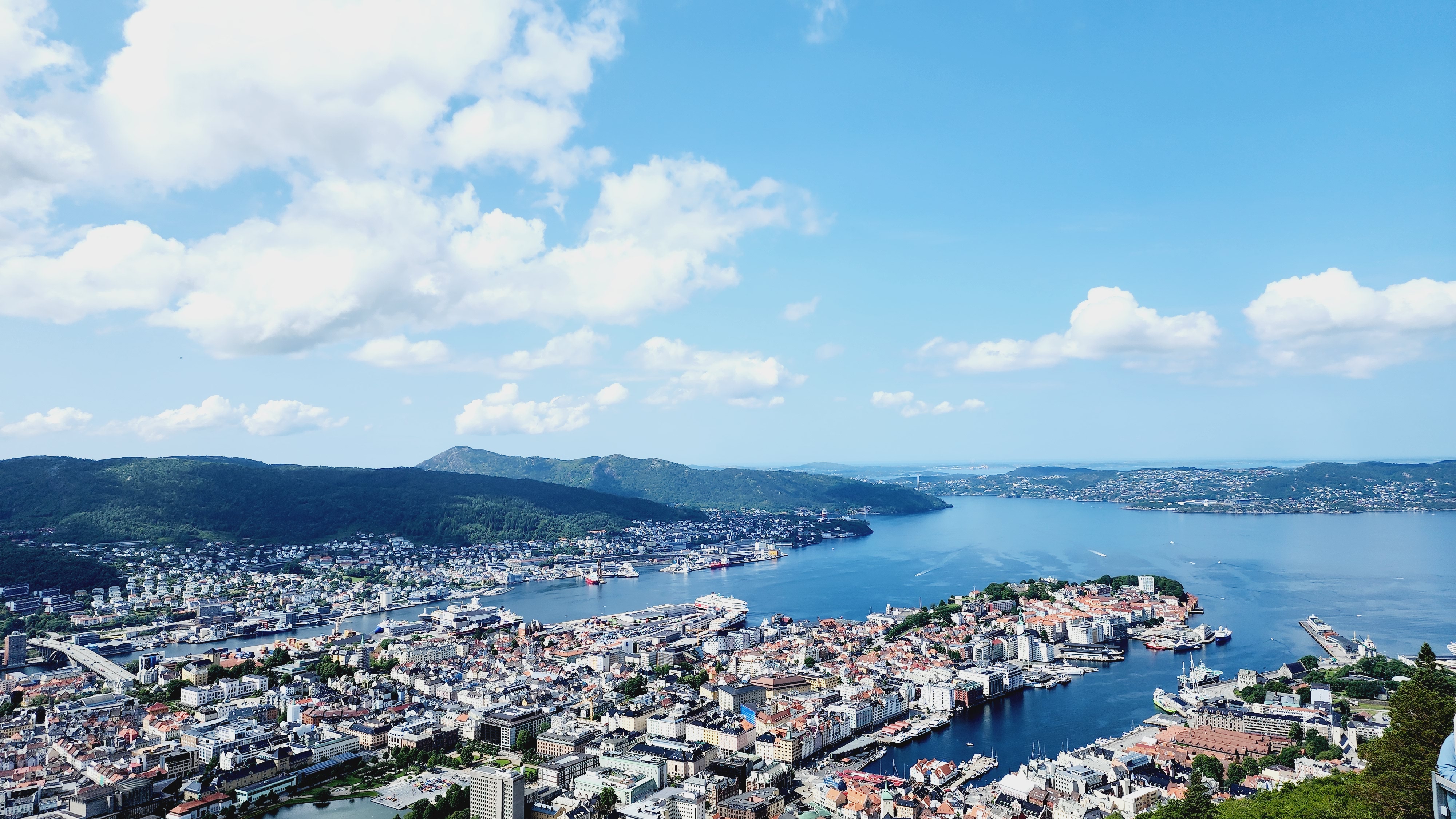 A panoramic view of Bergen, Norway, featuring its colorful buildings and bustling harbor nestled between verdant mountains. The calm blue waters of the fjord reflect the clear sky above, dotted with a few clouds. This scenic cityscape highlights Bergen's picturesque blend of natural beauty and urban charm.
