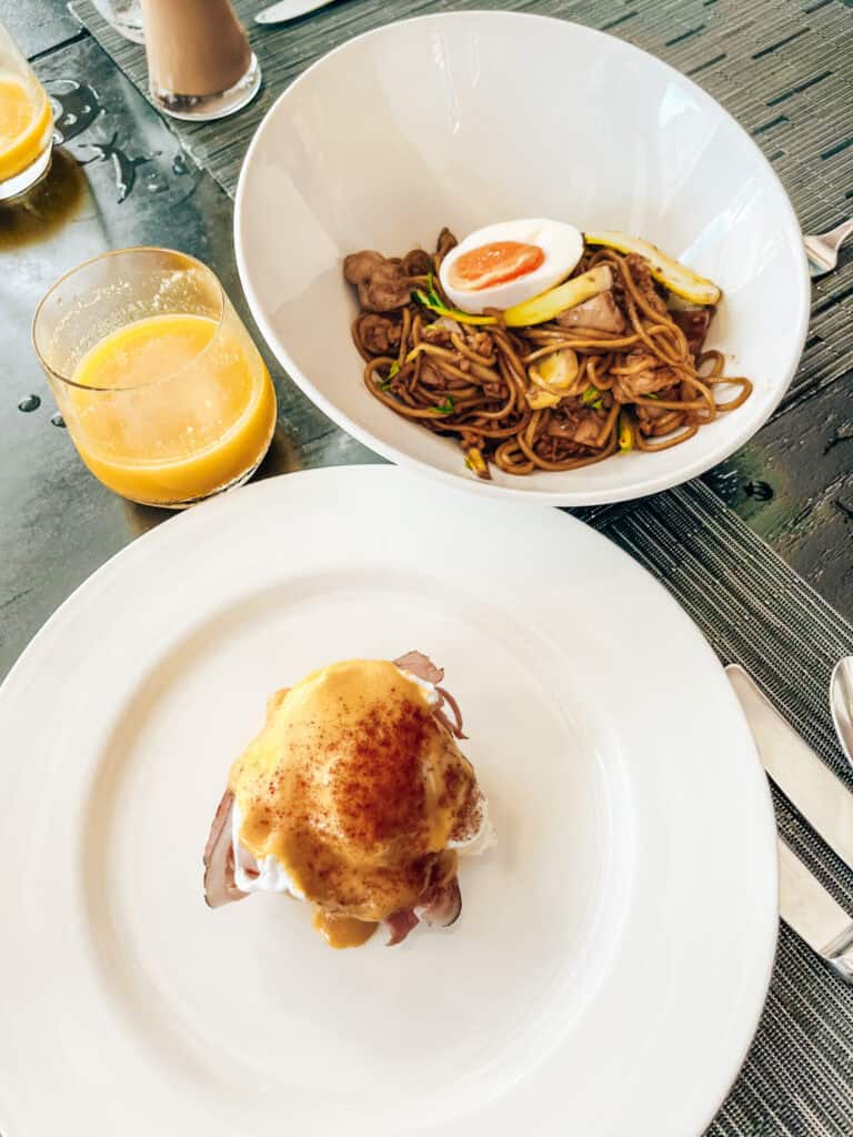 A breakfast setting featuring a bowl of noodles topped with a halved boiled egg, paired with a glass of orange juice, and a plate of eggs benedict drizzled with hollandaise sauce.
