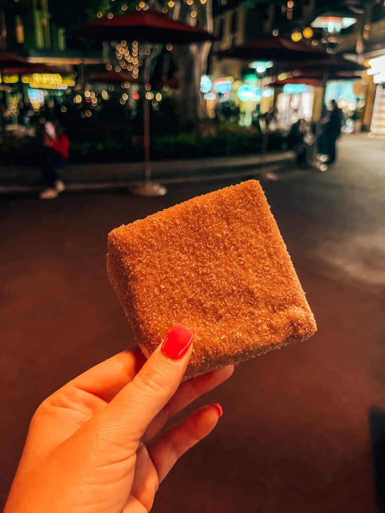 A hand with pink-painted nails holds a square piece of churro toffee, covered in cinnamon sugar, against a blurred night-time background with festive lights and people in the distance. The scene captures the warm glow of an evening out.