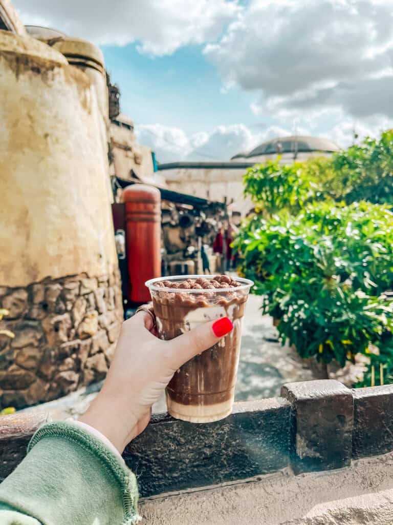 
A hand with red-painted nails holds a plastic cup of cold brew coffee topped with cream and cocoa puffs against a scenic, sunny background featuring themed architecture and greenery. The sky is partly cloudy, adding to the vibrant outdoor ambiance.