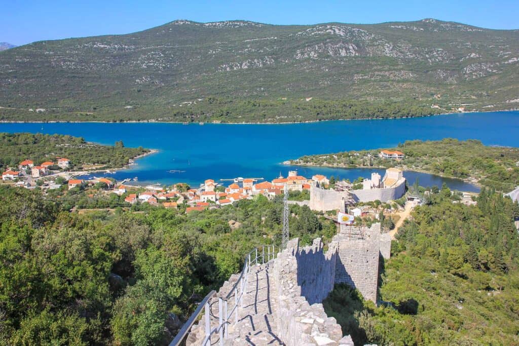 A scenic view of Dalmatia, Croatia, featuring a vibrant blue bay surrounded by green hills and dotted with small orange-roofed houses. The remains of an ancient stone wall and fortress stretch along the landscape, adding a historical element to the picturesque setting. The clear, sunny sky enhances the beauty of the coastal region.