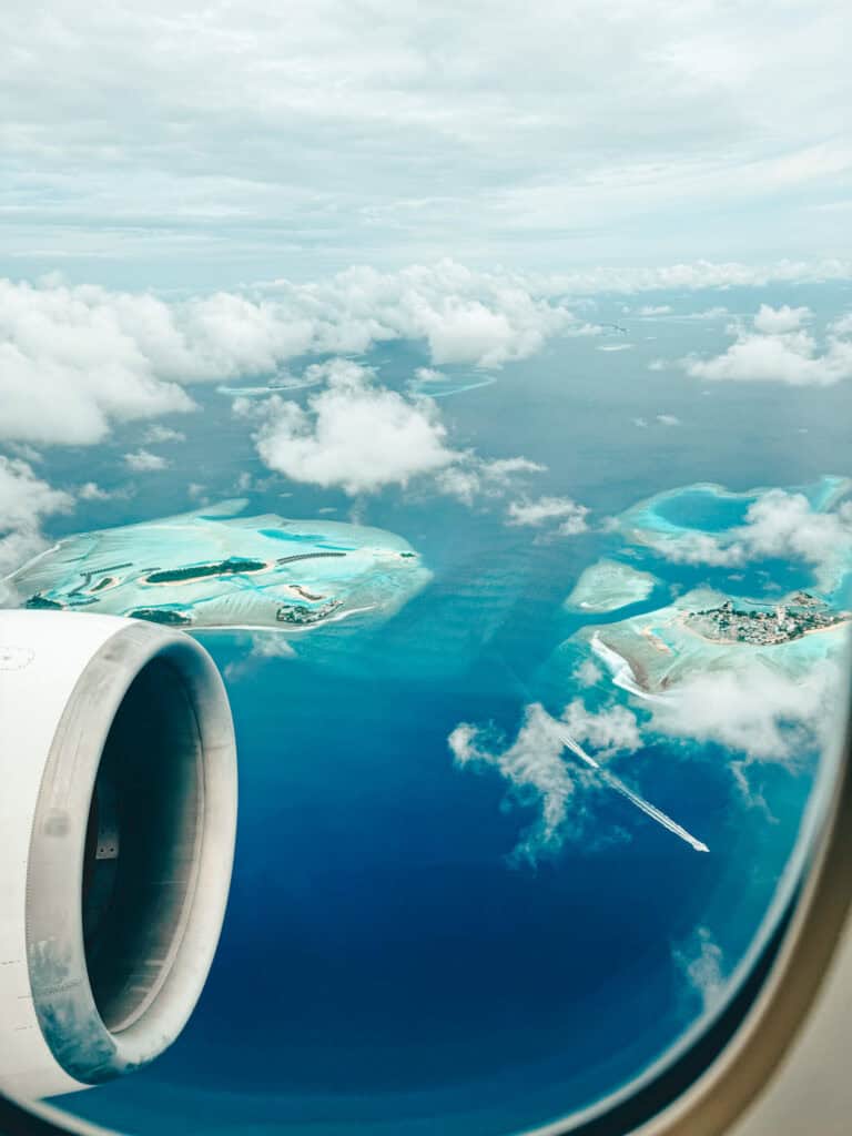 Aerial view from an airplane window showing the stunning blue waters of the Maldives, dotted with atolls and islands. The jet engine is visible on the left, framing a clear and expansive sky above and the deep blue ocean below, scattered with white clouds and tiny, vibrant islands