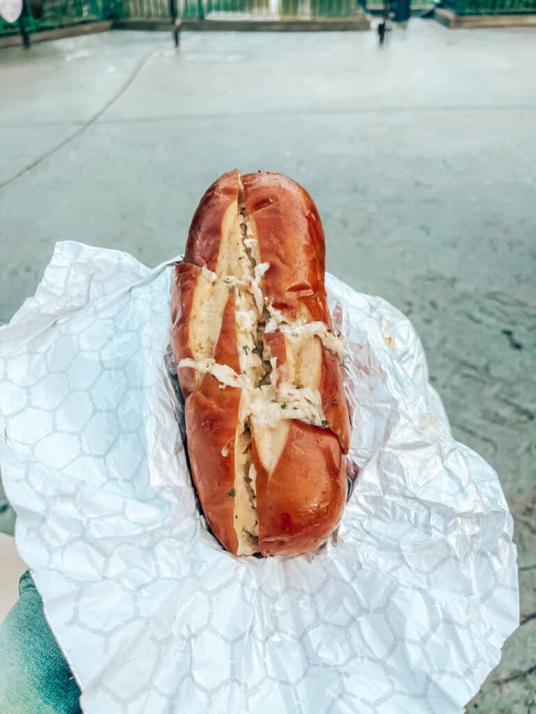 A hand holds a garlic butter-filled pretzel bun wrapped in white wax paper. The background features a blurred outdoor setting with a paved ground and green fencing.