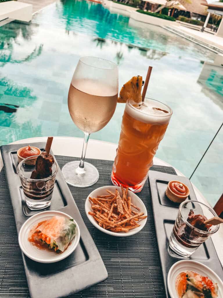 An assortment of appetizers including spring rolls, seasoned fries, and various small bites, accompanied by a glass of chilled rosé and a tropical cocktail with a pineapple garnish, set beside a tranquil pool.
