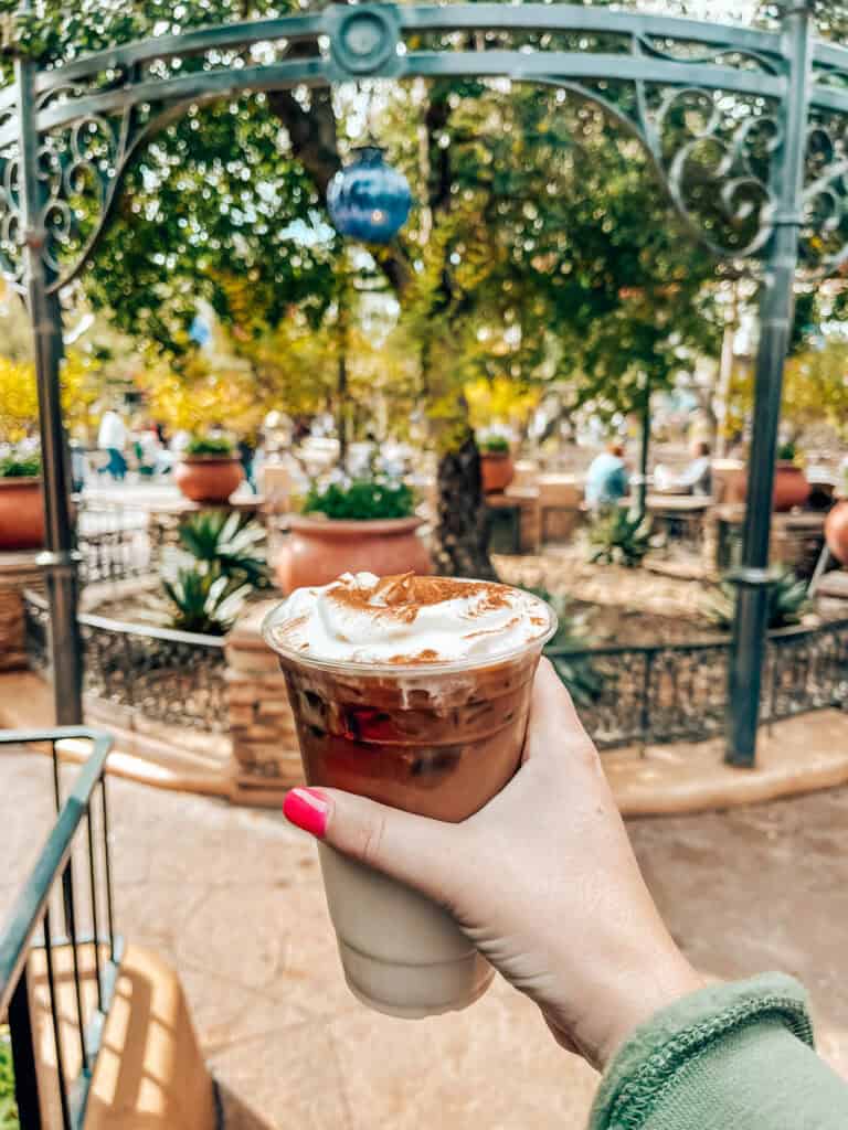 A hand with pink-painted nails holds a plastic cup of horchata cold brew topped with whipped cream and a sprinkle of cinnamon. The background features a lush, outdoor seating area with decorative wrought iron and potted plants, creating a cozy and inviting atmosphere.