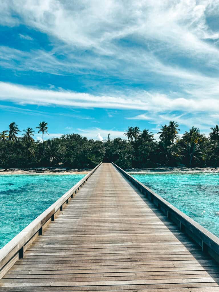 Long wooden jetty extending towards a lush tropical island in the Maldives. The jetty is flanked by crystal clear turquoise waters under a bright blue sky adorned with wispy clouds, leading the viewer's eye towards the dense greenery of palm trees on the island.