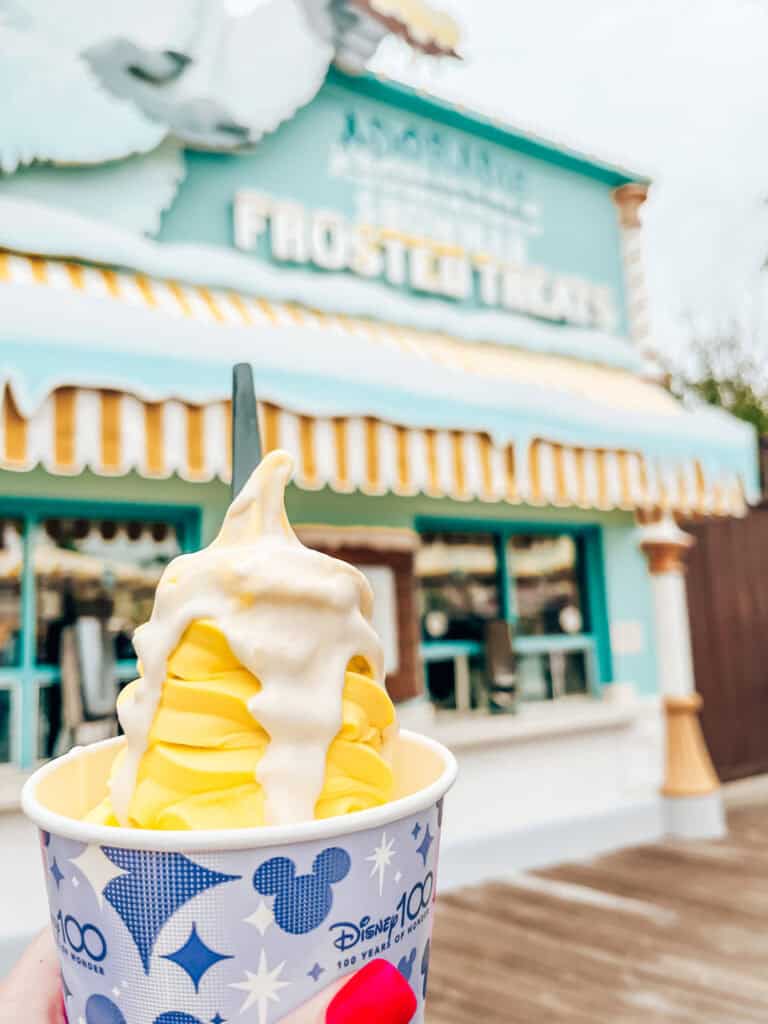 
A hand with pink-painted nails holds a cup of lemon soft serve topped with creamy white sauce, in front of Adorable Snowman Frosted Treats. The background features the shop's colorful signage and festive décor, creating a cheerful and inviting atmosphere.